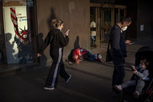 A man sleeps on the sidewalk while pedestrians walk by and look at their smartphones in downtown Barcelona.